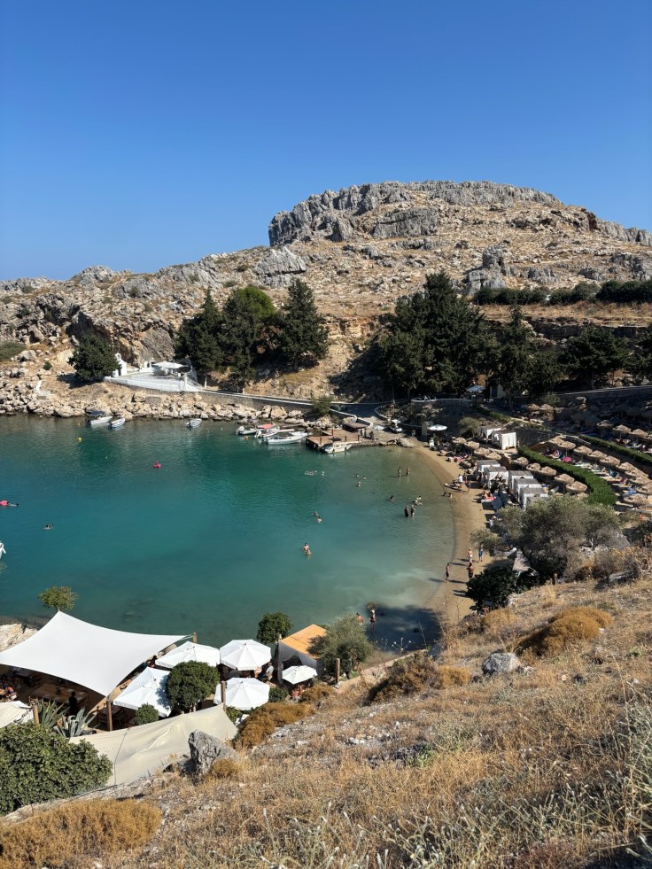 Close‑up view of St Paul’s Bay with turquoise water, sandy beach, and rocky cliffs in Lindos, Rhodes.