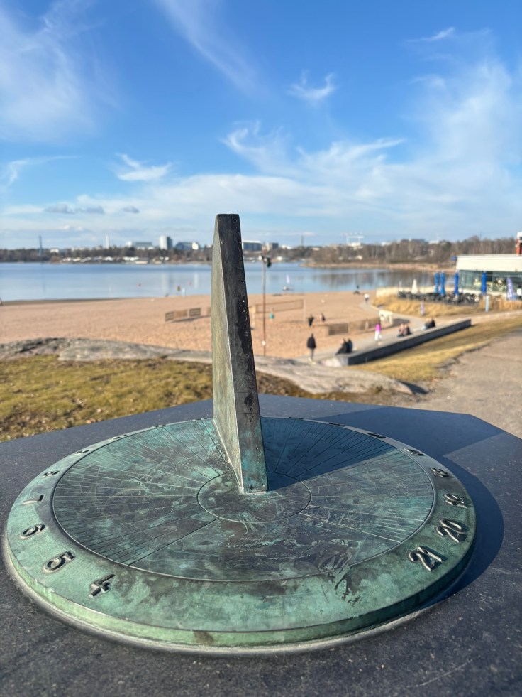 Sundial overlooking a sandy beach and calm waterfront in Helsinki, Finland under a clear blue sky