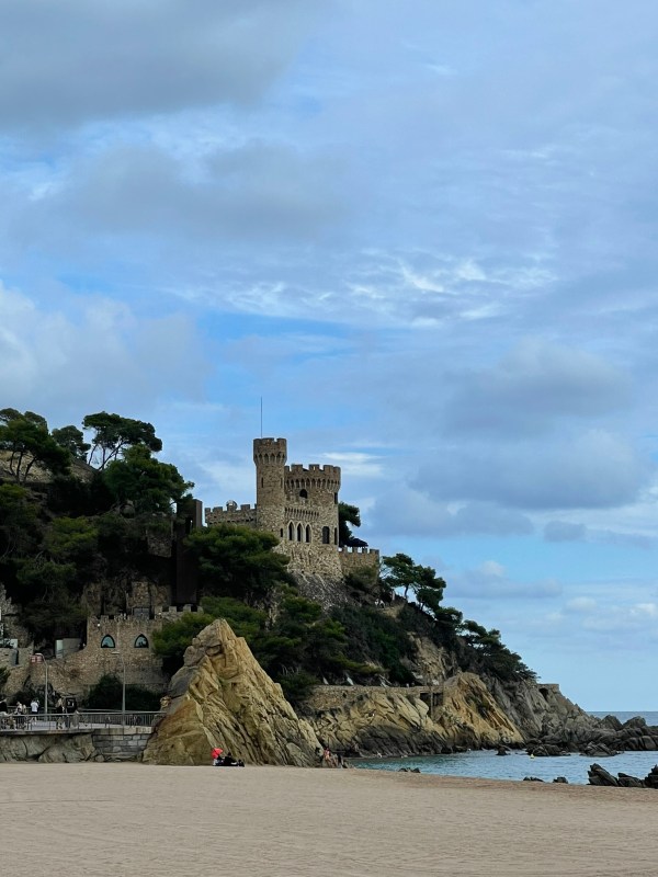 Castle perched on a rocky cliff above a sandy beach under dramatic cloudy skies